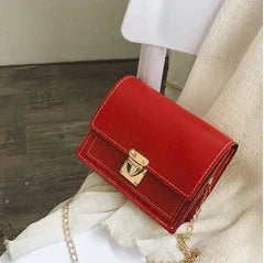 Small red leather crossbody bag with gold clasp and chain strap on white chair