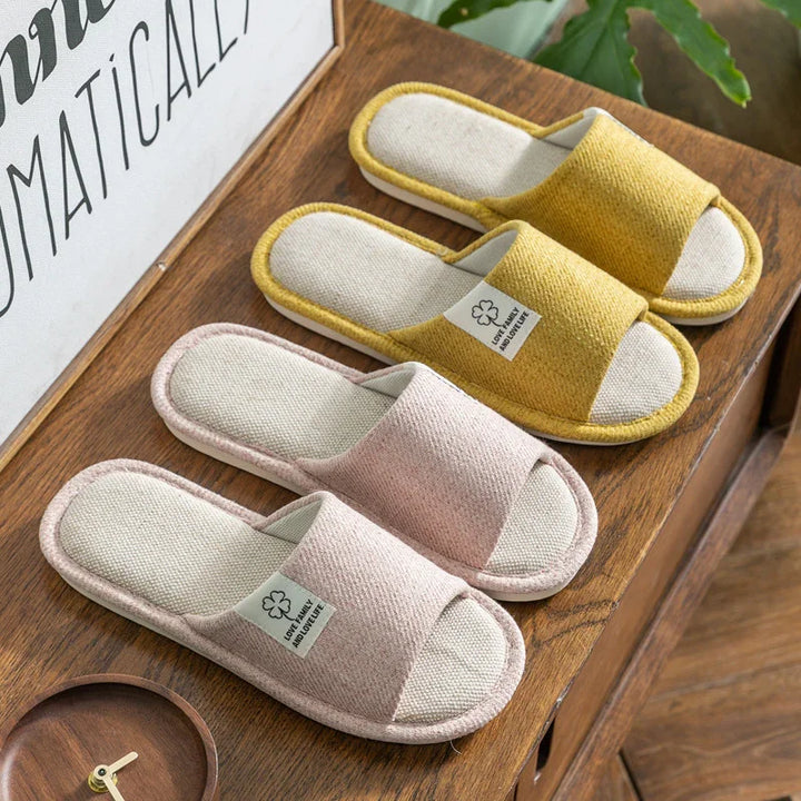 Two pairs of open-toe fabric slippers in pink and yellow on wooden table with green plant