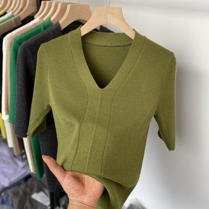 Hand holding olive green ribbed knit V-neck short sleeve sweater on wooden hanger with assorted sweaters in background