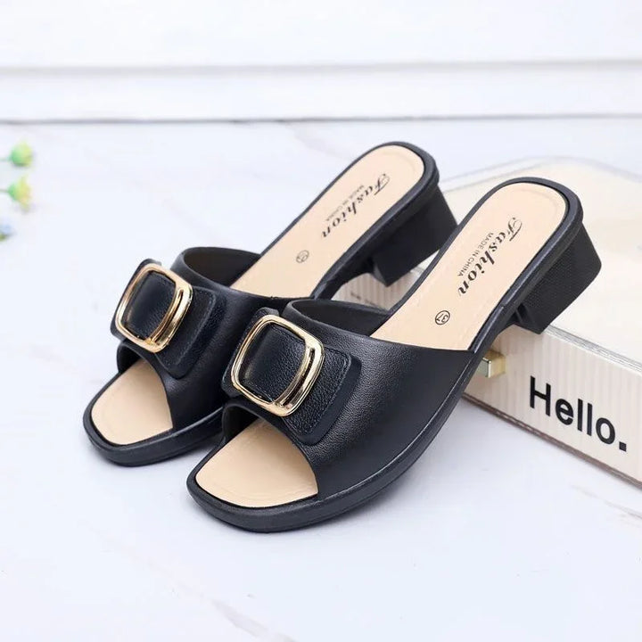Black open-toe block heel sandals with gold buckle detail on white surface next to a box labeled Hello