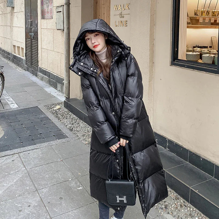 Woman wearing long black hooded puffer coat and holding black handbag on urban sidewalk