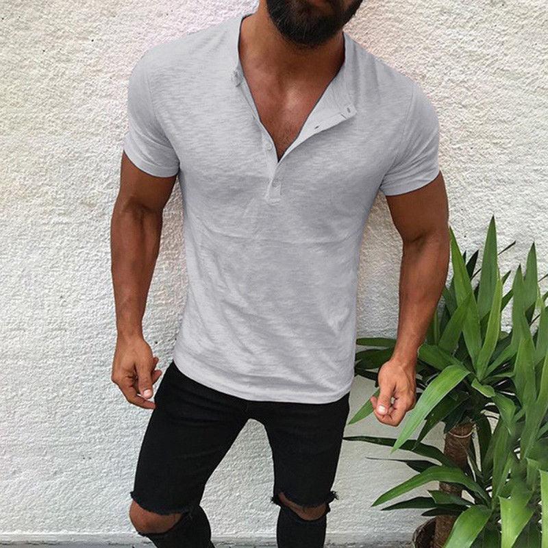 Muscular man wearing a fitted light grey henley t-shirt and ripped black pants, standing against white wall with green plant