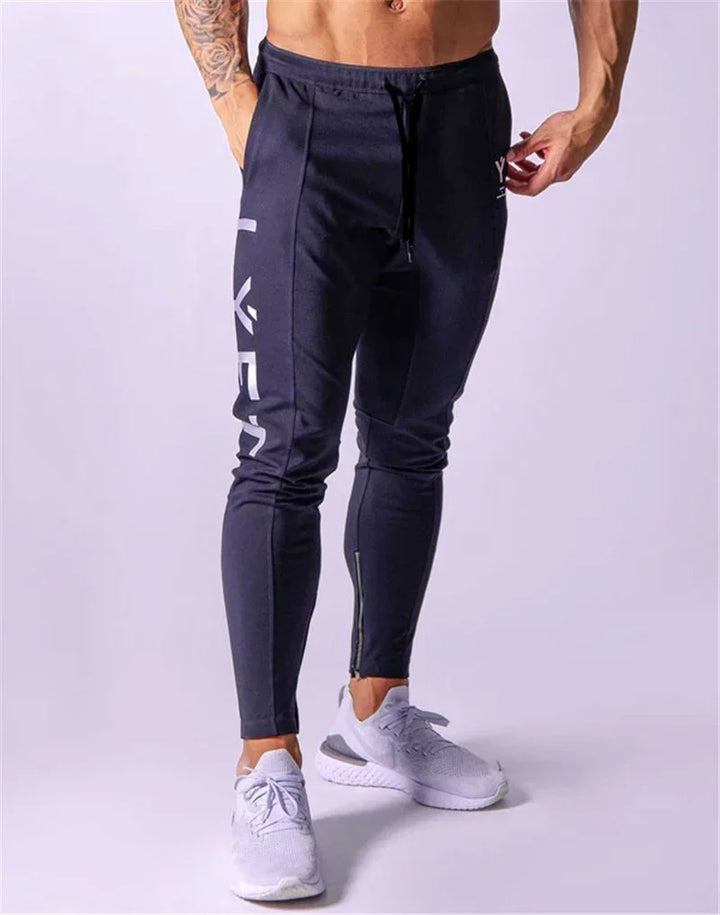 Male model wearing slim fit black gym joggers with side print and white sneakers on light background