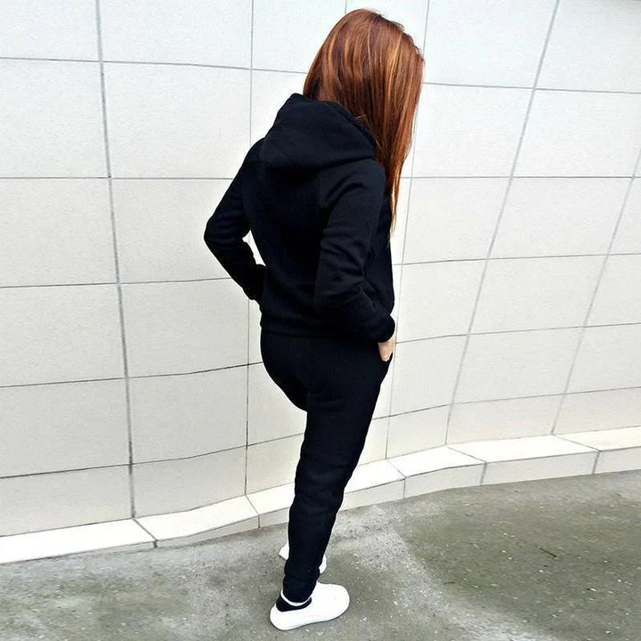 Person with red hair wearing black hoodie and sweatpants, white sneakers standing against tiled wall