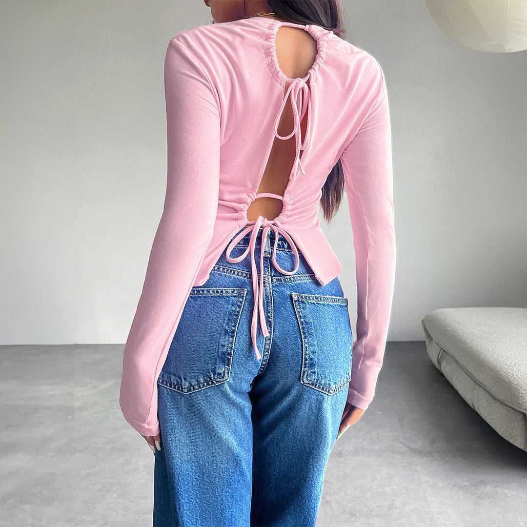 Woman wearing a pink open-back long sleeve top with tie details paired with blue denim jeans indoors