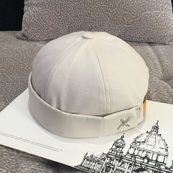 Beige cotton docker cap with a rolled brim and metal crossed pins, placed on an architectural drawing