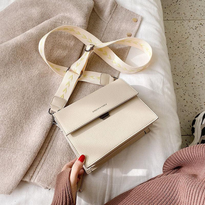 Beige leather crossbody bag with patterned strap resting on a white bed beside a beige coat and a person wearing a brown sweater