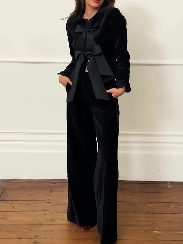 Woman wearing black velvet wide-leg pants and matching jacket with large bows, standing on wooden floor