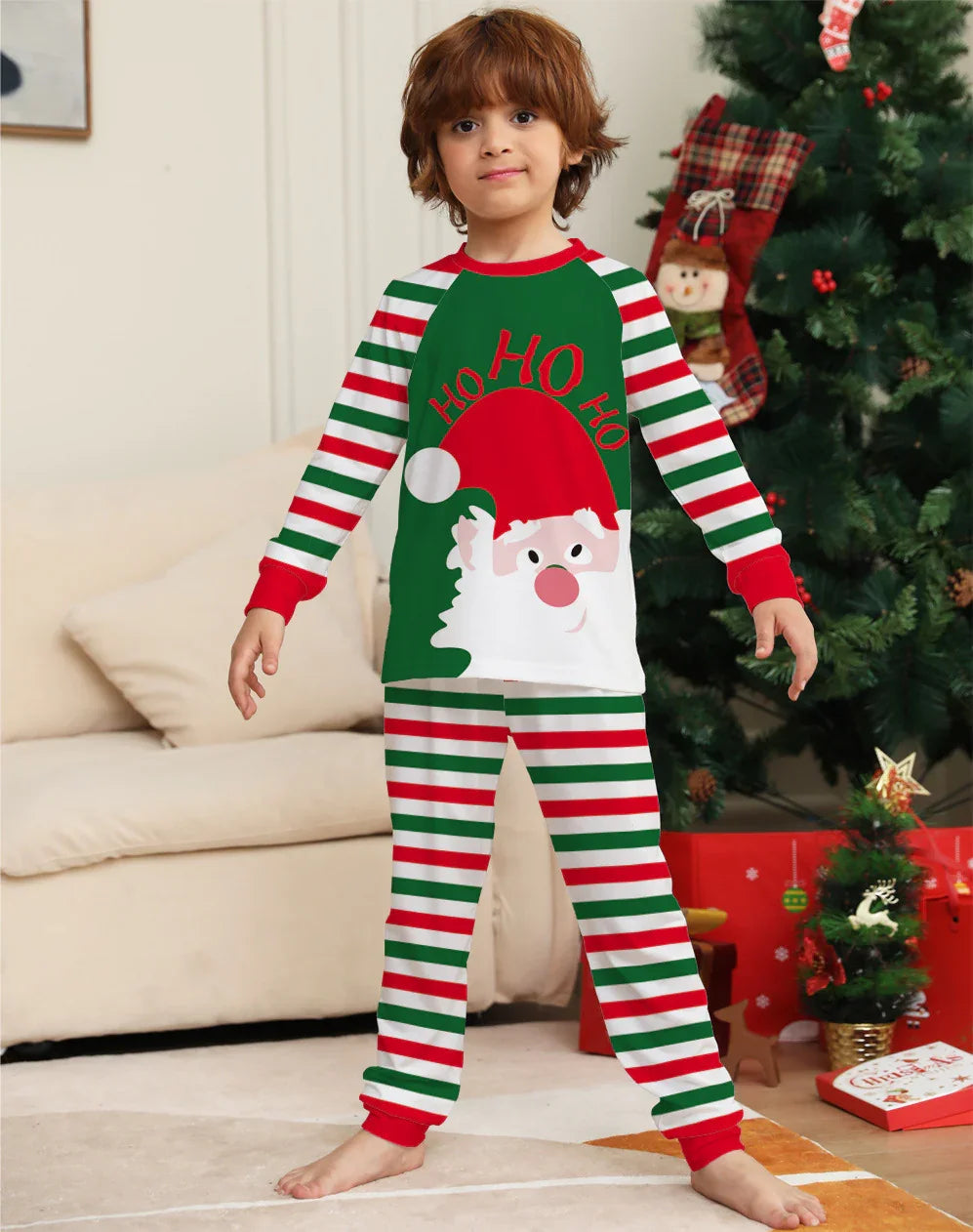 Child in Christmas pajamas with Santa and 'Ho Ho Ho' design, standing near decorated tree and presents
