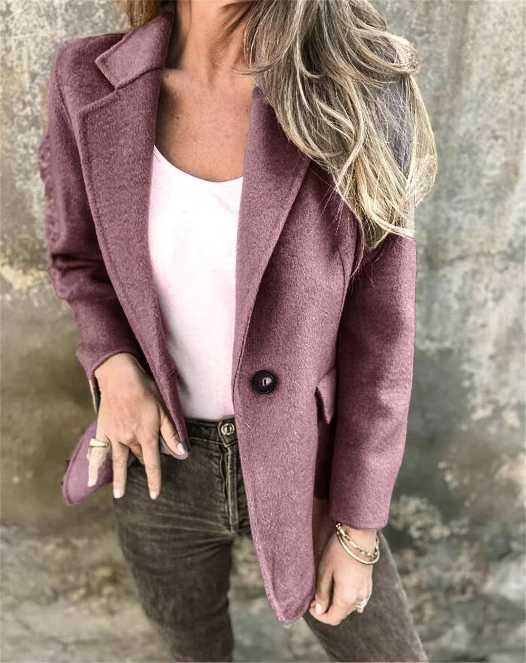 Woman wearing dusty rose blazer over white top and brown pants standing against textured wall