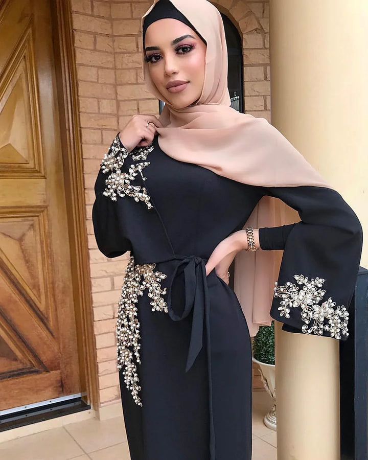 Woman in black abaya with gold floral embroidery and beige hijab standing near wooden door