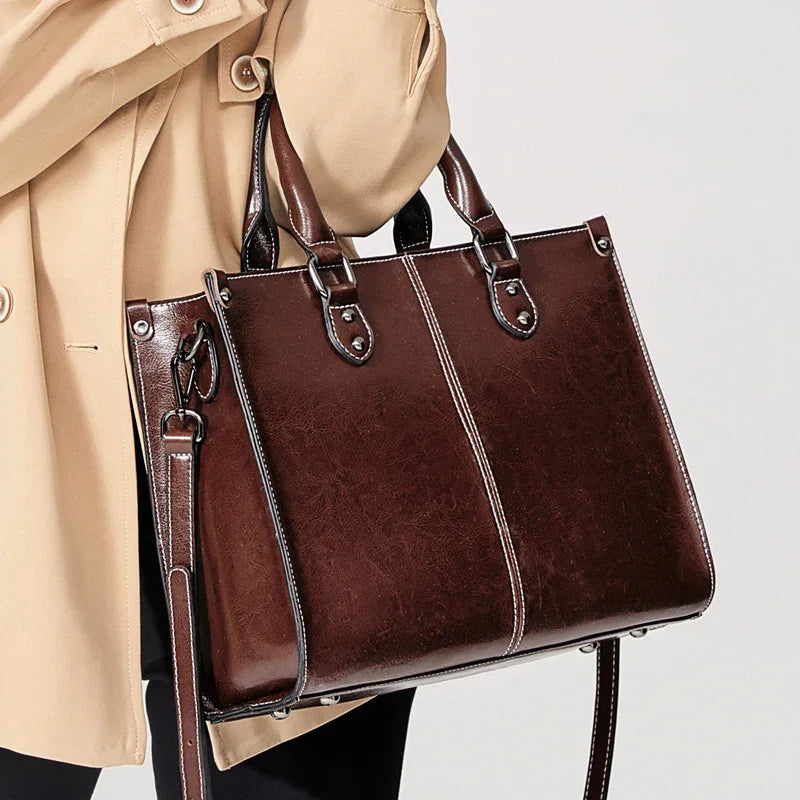 Brown leather handbag with white stitching and metal details carried by a person wearing a beige coat