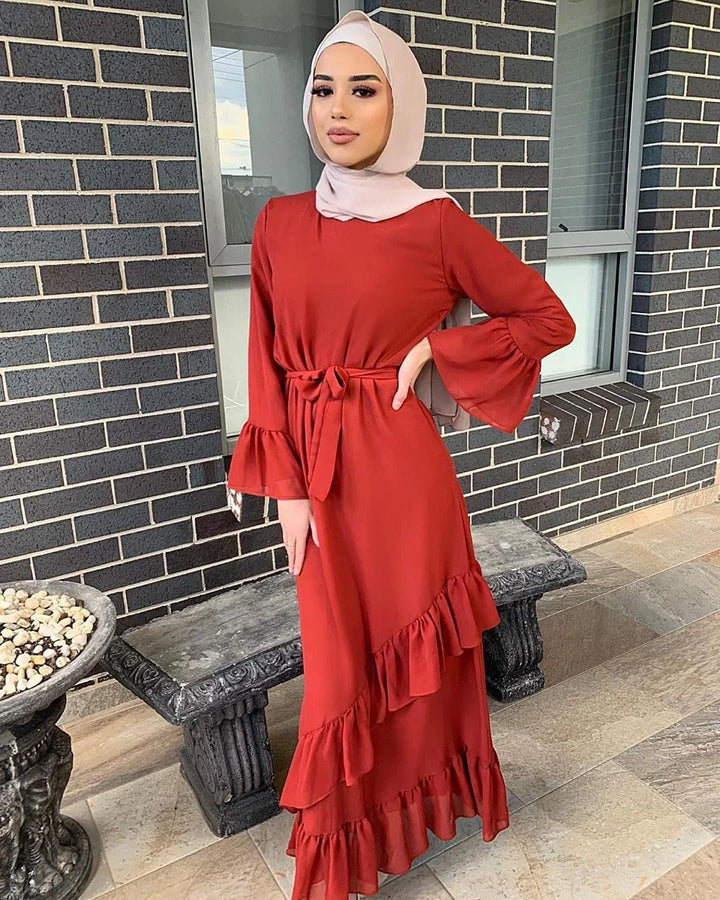 Woman wearing modest red ruffle dress and beige hijab standing by brick wall and stone bench
