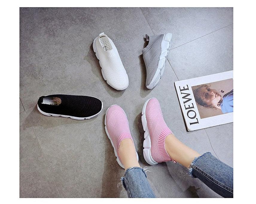 Pink knit slip-on sneakers on feet surrounded by white, gray, and black knit slip-on shoes on tiled floor