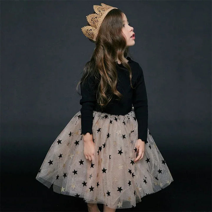 Young girl wearing a black top, star-patterned tulle skirt, and gold crown against a dark background