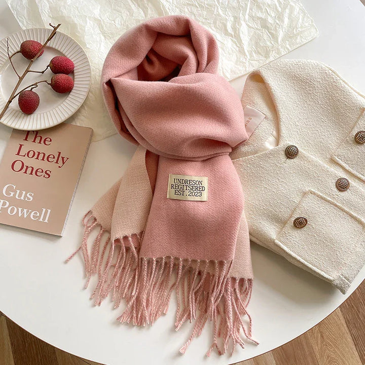 Soft pink fringed scarf with label, cream textured jacket with buttons, book, and lychee fruit on table