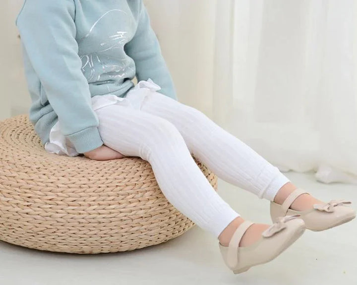 Child wearing white ribbed leggings and beige bow shoes sitting on woven pouf ottoman