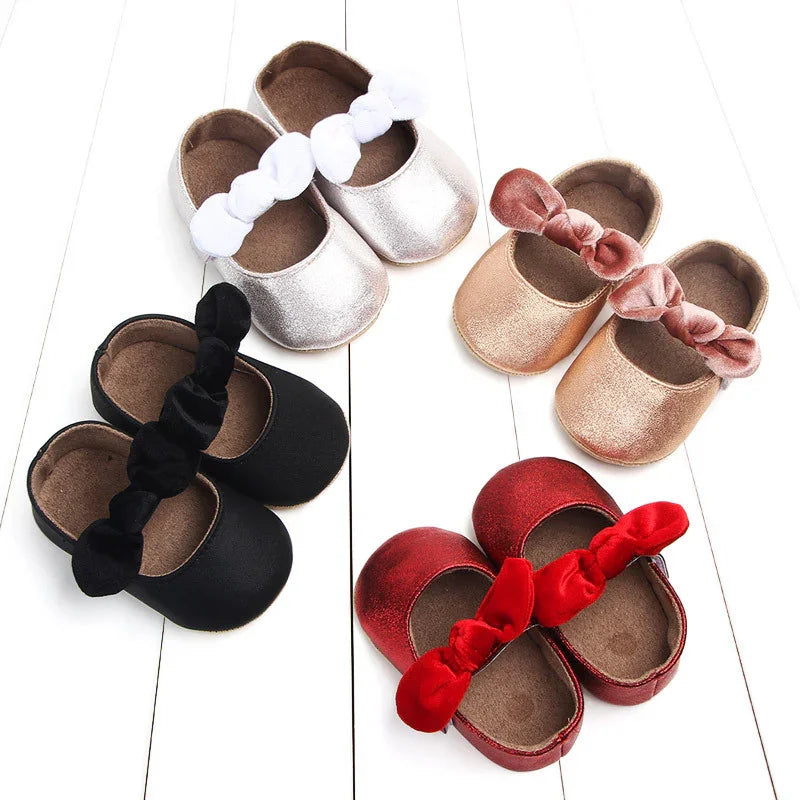 Four pairs of baby shoes in black, white, rose gold, and red with matching velvet bows on white wooden floor