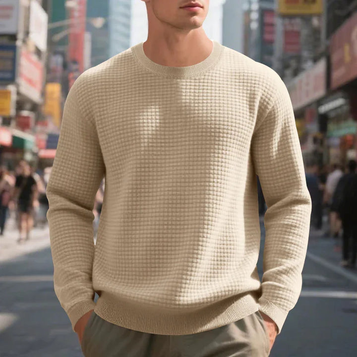 Man wearing beige textured long-sleeve sweater in urban street setting with blurred pedestrians