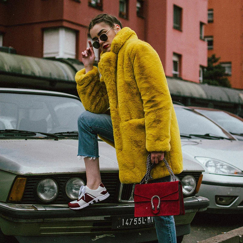 fashionable woman in yellow faux fur coat and sunglasses posing on vintage car with red handbag and jeans