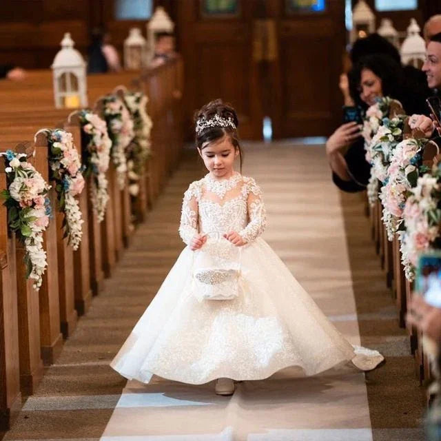 Young girl in white lace dress and tiara walking down flower-decorated aisle at wedding
