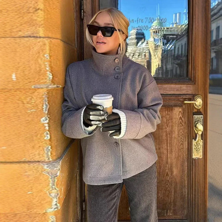 Woman wearing gray coat, black gloves, and sunglasses holding a coffee cup by a wooden door