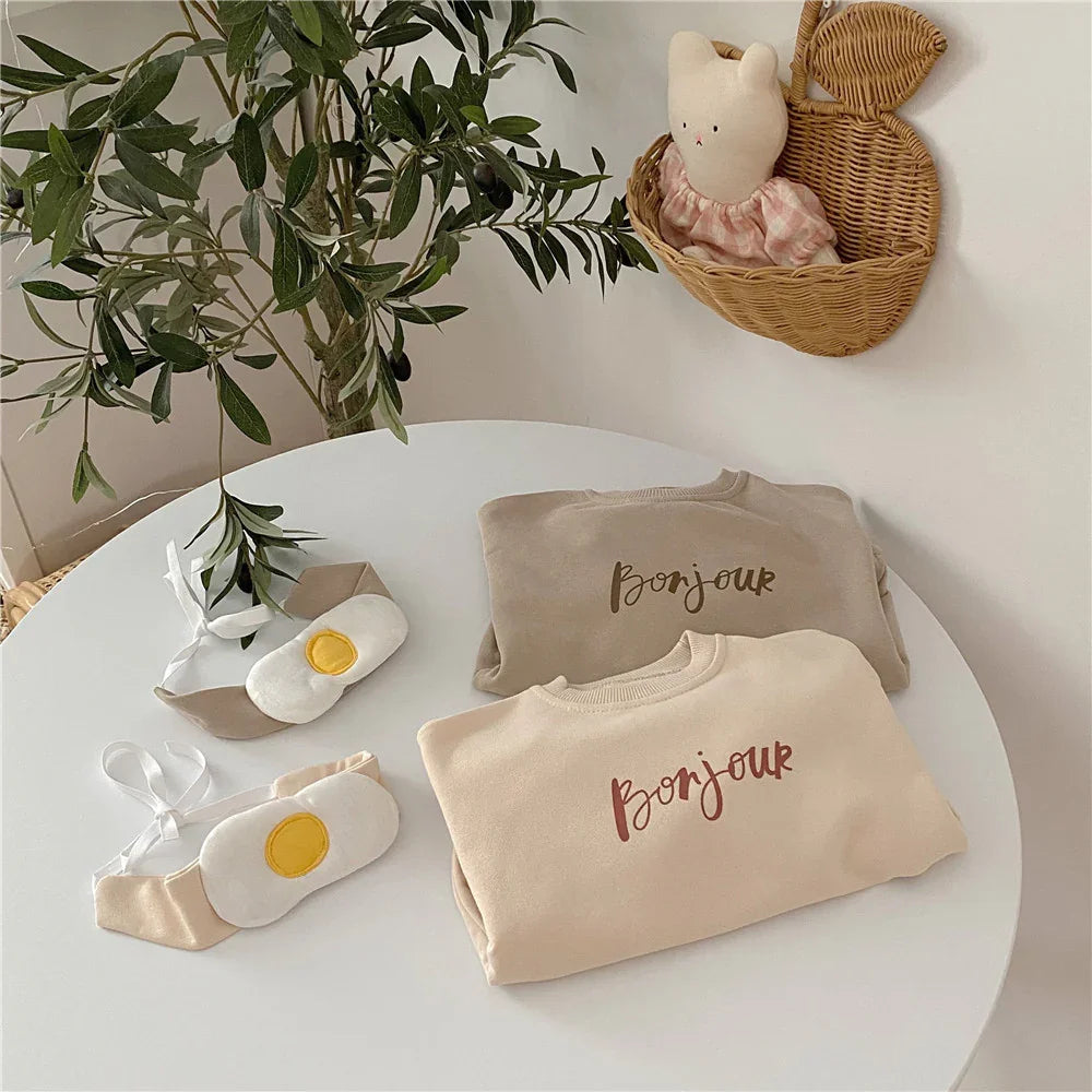 Two folded beige children's sweatshirts with "Bonjour" text on white round table with egg-themed baby shoes and hanging wicker basket with plush toy