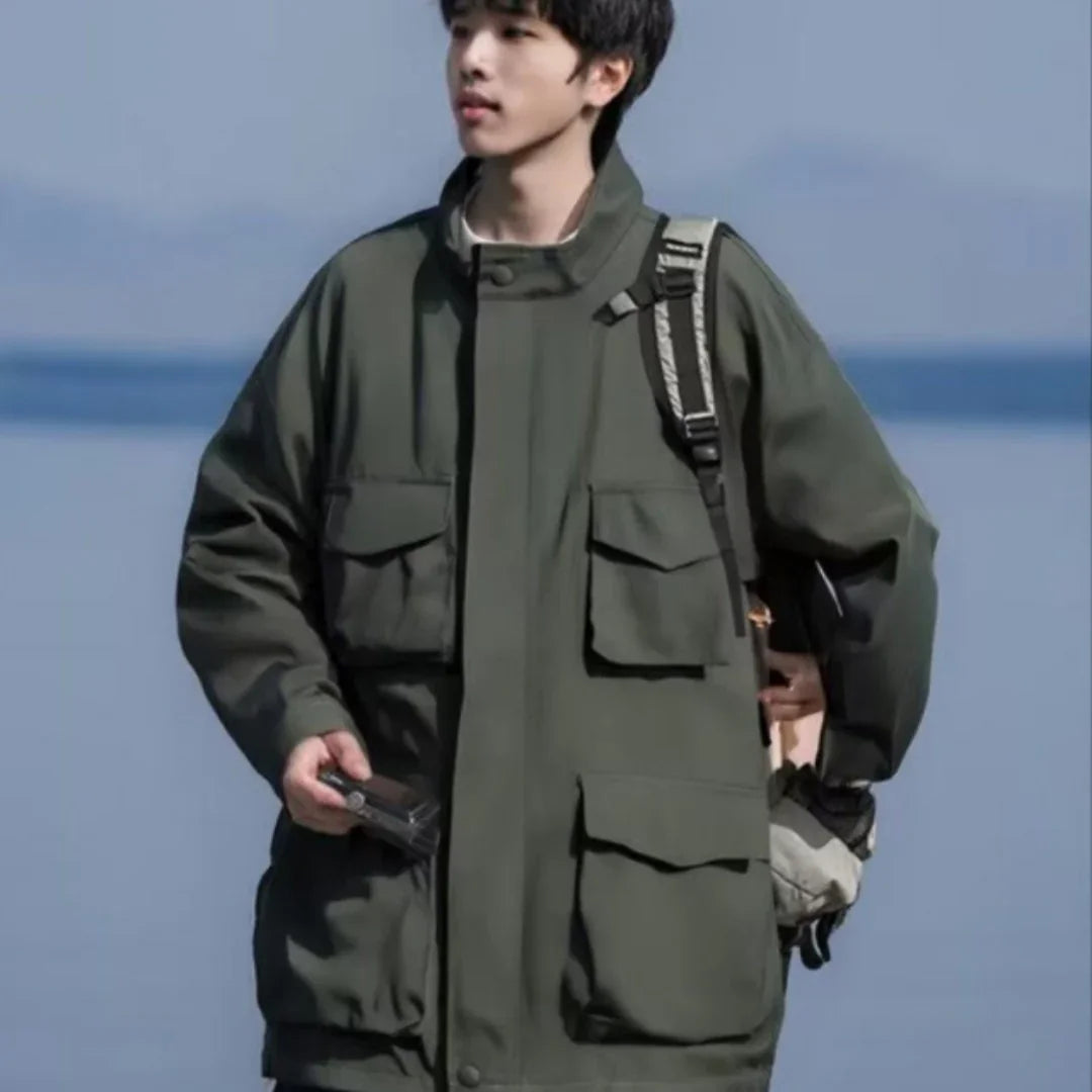 Young man wearing olive green utility jacket and carrying backpack, standing outdoors by water