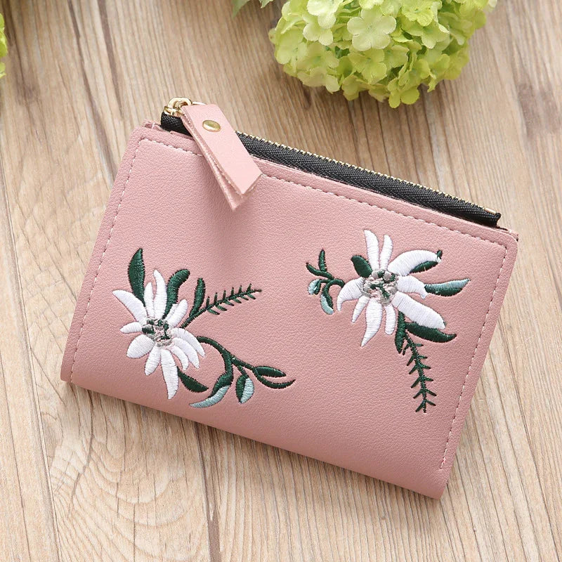 Pink leather wallet with white floral embroidery and zipper on wooden surface
