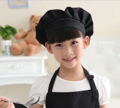Young girl wearing black chef hat and apron holding a spoon in a bright kitchen