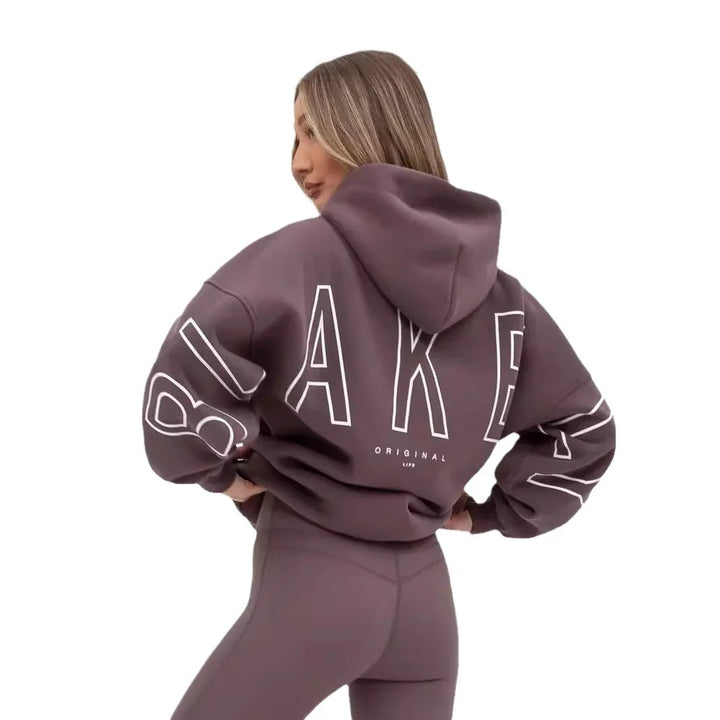 Woman wearing a mauve oversized hoodie with 'BAKER' print and matching leggings, posing against white background