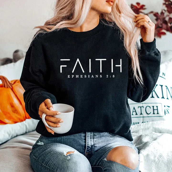 Woman wearing black sweatshirt with 'Faith Ephesians 2:8' text, holding white mug, sitting on couch