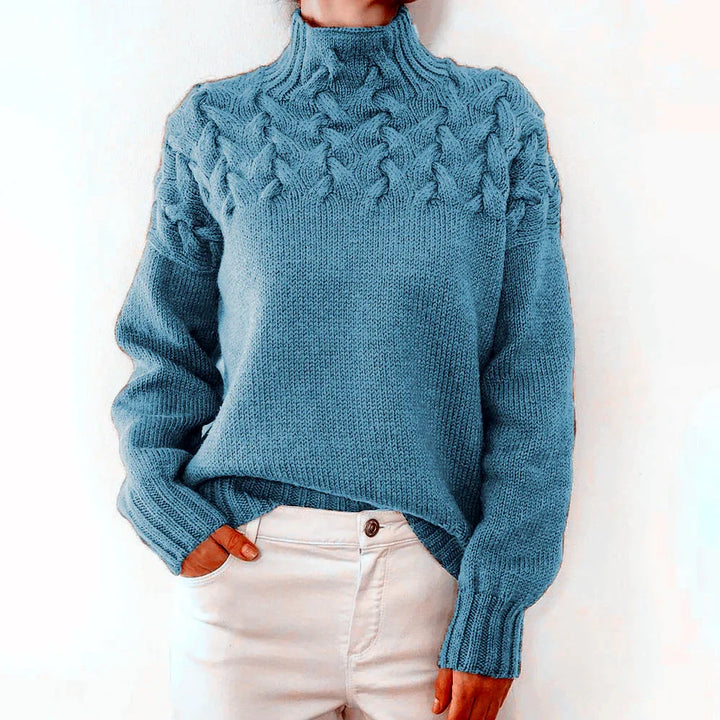 Person wearing a blue cable knit turtleneck sweater and white pants against a white background