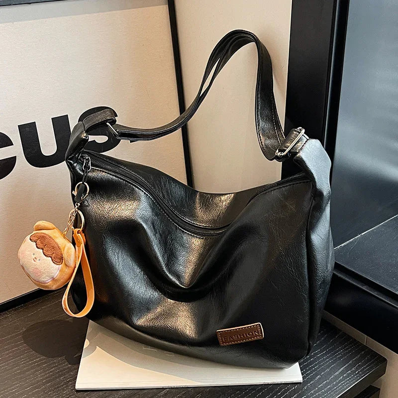 Black leather shoulder bag with adjustable strap and cute plush keychain on a dark wooden table