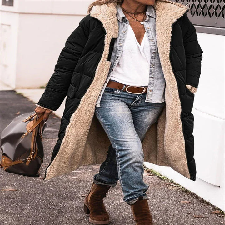 Woman walking outdoors wearing long black puffer coat with beige sherpa lining, blue jeans, brown boots, and carrying brown handbag