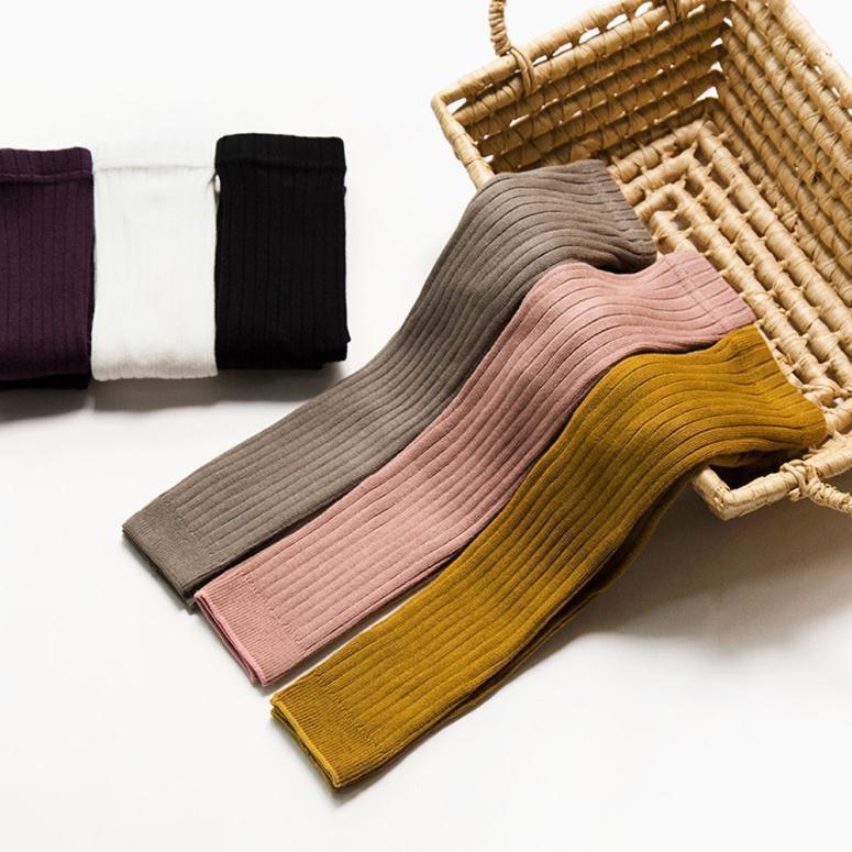 Ribbed knit leggings in mustard, pink, and gray draped over a wicker basket with folded socks in purple, white, and black nearby