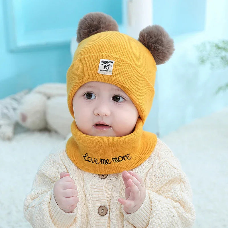 baby wearing yellow knit hat with pom poms and matching neck warmer on cozy background