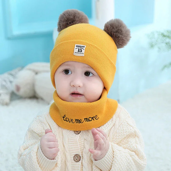 baby wearing yellow knit hat with pom poms and matching neck warmer on cozy background
