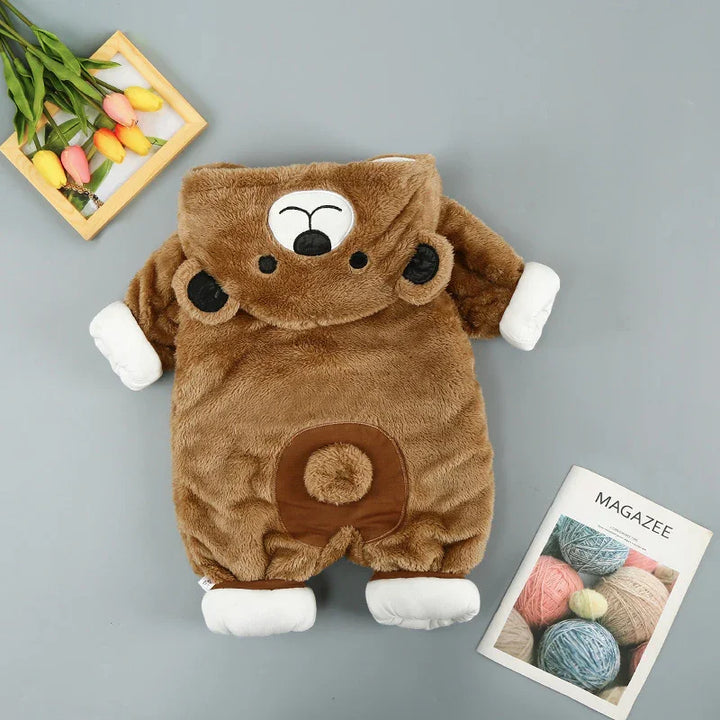 Brown plush baby romper with bear face and ears on hood, white cuffs, laid flat on gray surface