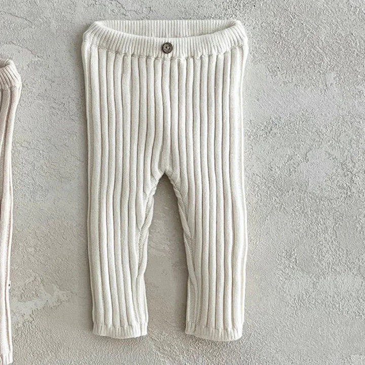 Cream ribbed knit baby pants with button detail on waist, displayed on textured gray background