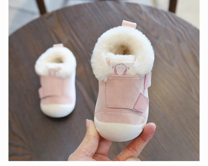Hand holding a soft pink baby shoe with white faux fur lining, placed on a wooden table with matching shoe in background