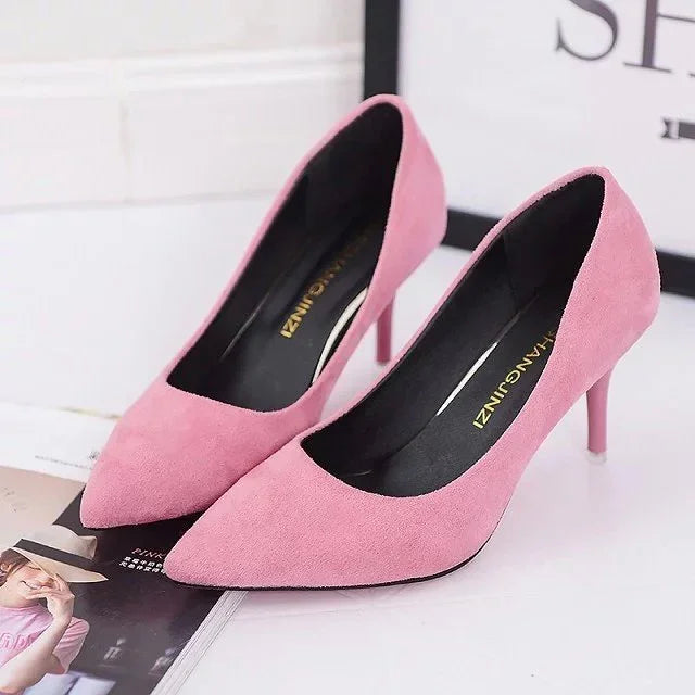 Pair of elegant pink suede pointed-toe high heel shoes on white surface with magazine and frame background