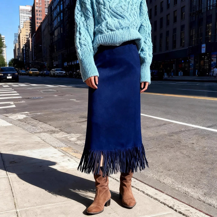 Model wearing a blue fringed midi skirt, light blue cable knit sweater, and brown suede boots on city sidewalk