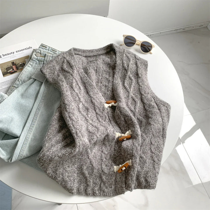 Gray cable knit wool vest with toggle buttons on white round table with light blue jeans and beige sunglasses