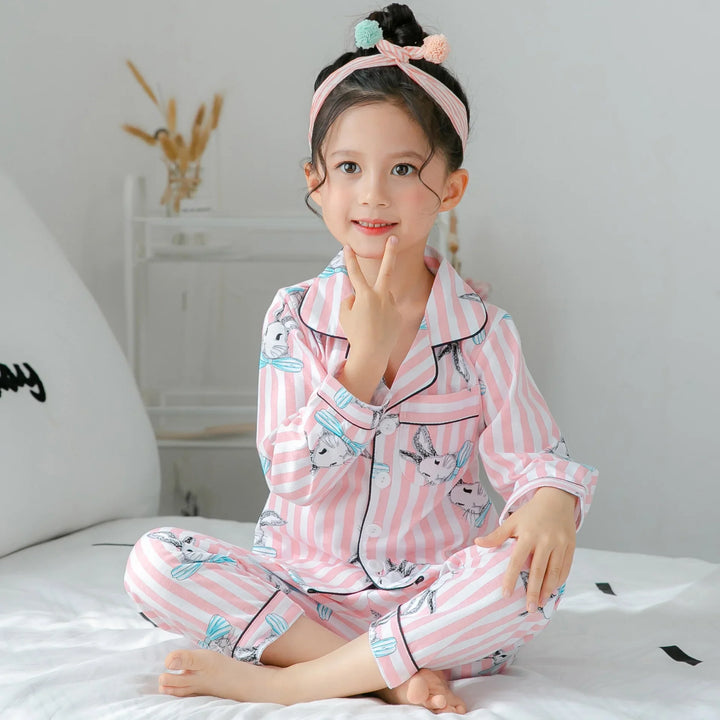 Girl in pink striped bunny print pajamas and floral headband sitting cross-legged on white bed