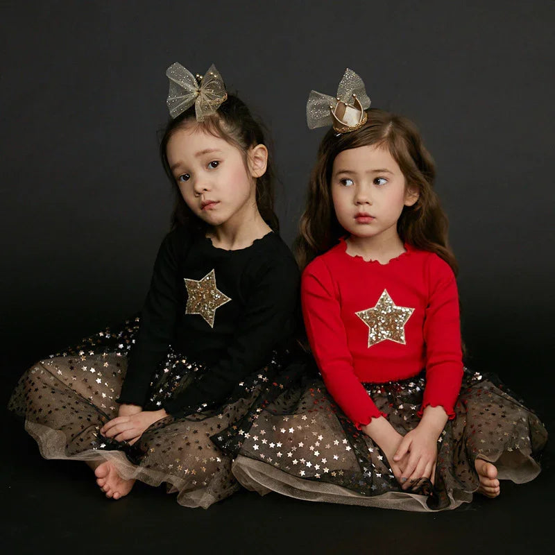 Two young girls in star-themed outfits with tulle skirts, black and red tops, and glittery bows against black background