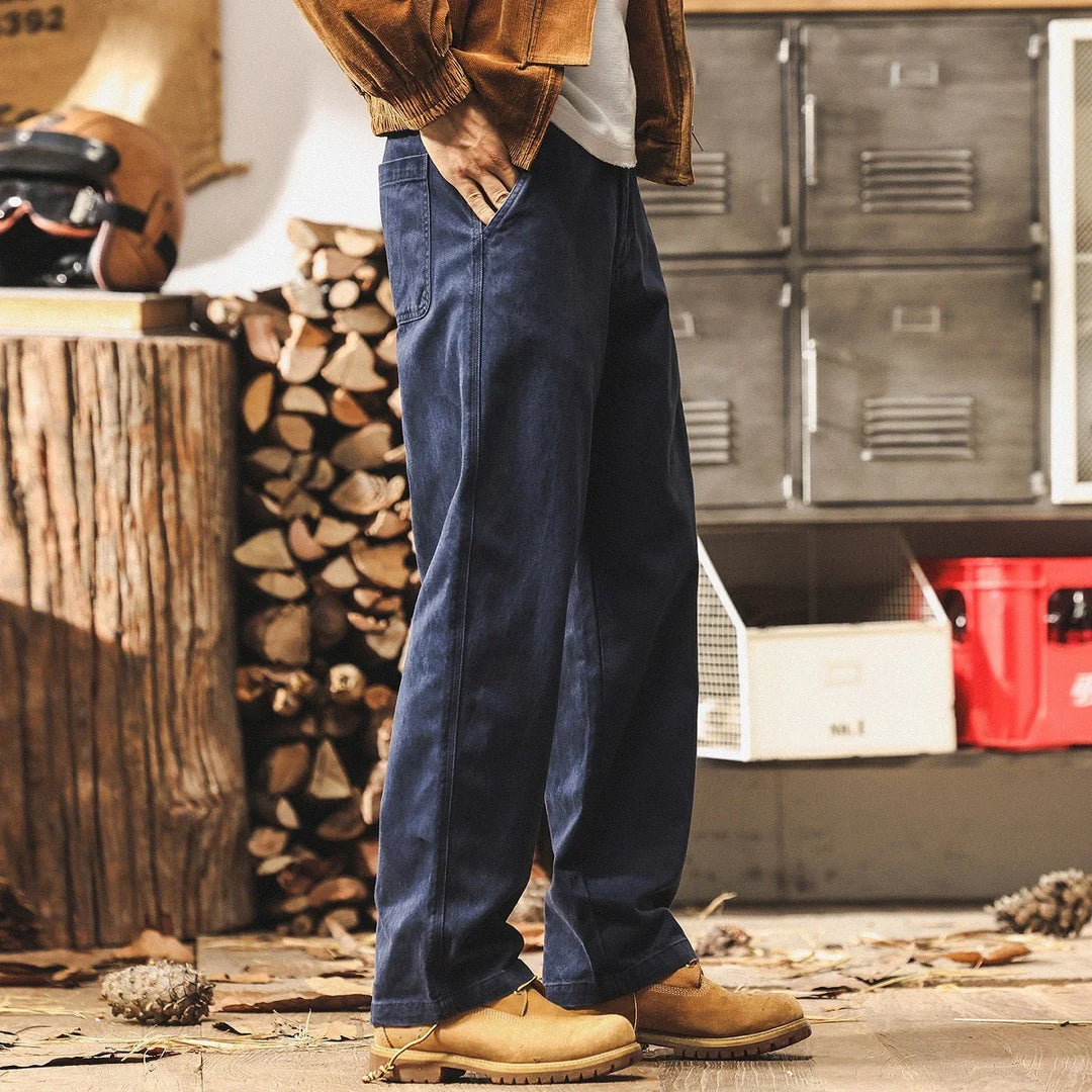 Man wearing relaxed fit navy blue pants and tan boots in rustic indoor setting with firewood and metal lockers