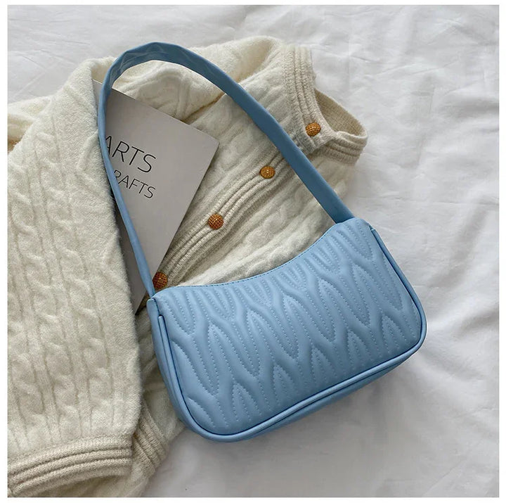 Light blue textured shoulder bag placed on cream knit sweater with gold buttons and arts crafts book