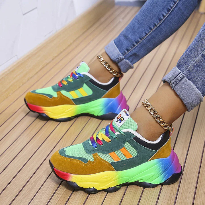 Colorful rainbow chunky sneakers with green, orange, and brown panels worn with rolled-up jeans and gold chain anklets on wooden floor
