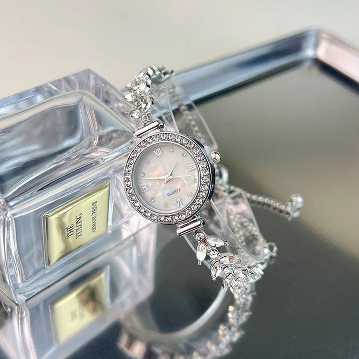Silver quartz wristwatch with crystal bezel and leaf bracelet on reflective surface next to perfume bottle